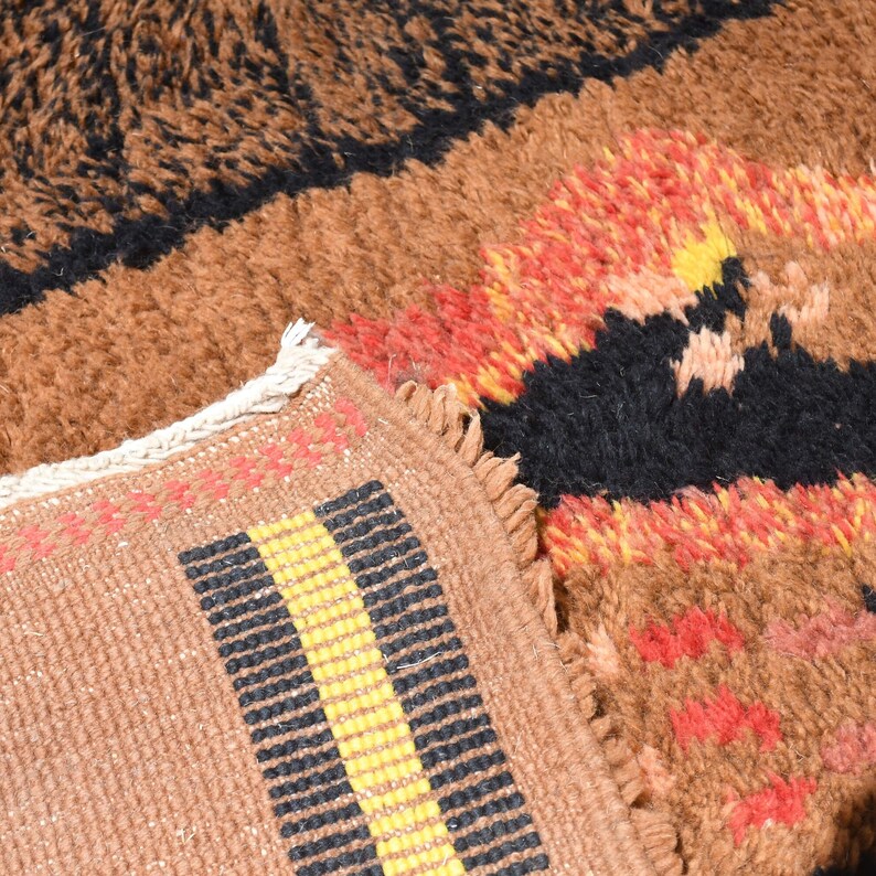 May include: Close-up of a brown and black patterned rug with a yellow, black, and red stripe detail. The rug has a fuzzy texture and a fringe border.