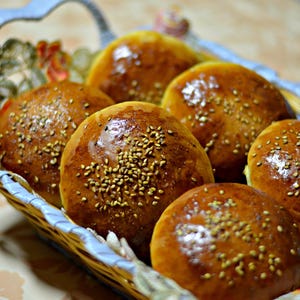 May include: A basket holds six golden-brown, sesame-topped buns. The buns have a glossy finish, suggesting they are freshly baked. The basket has a decorative floral lining and a light blue handle.