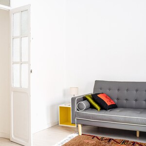 May include: A brown and gray patterned rug with colorful accents, placed in front of a gray sofa with pillows. The sofa is in a white room with a white door and a yellow side table.