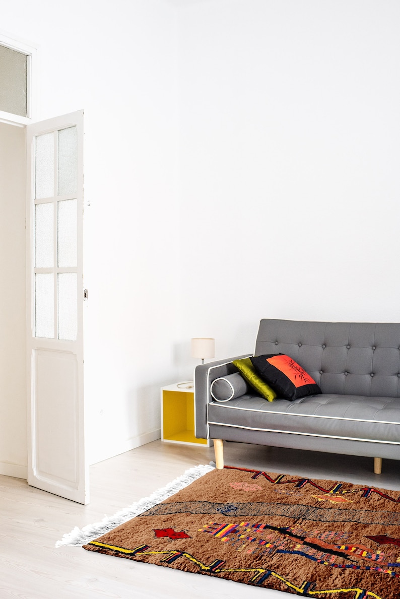 May include: A brown and gray patterned rug with colorful accents, placed in front of a gray sofa with pillows. The sofa is in a white room with a white door and a yellow side table.