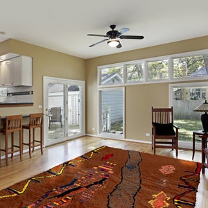 May include: A large, brown area rug with a colorful, abstract design. The rug is laid out in a living room with two wooden chairs and a table with a lamp.