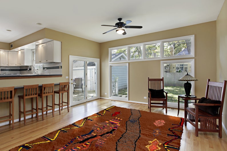 May include: A large, brown area rug with a colorful, abstract design. The rug is laid out in a living room with two wooden chairs and a table with a lamp.