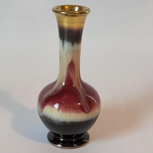 May include: A small, ceramic vase with a rounded body and a narrow neck. The vase is decorated with a multi-colored glaze in shades of red, white, and black. The top of the vase has a gold rim.