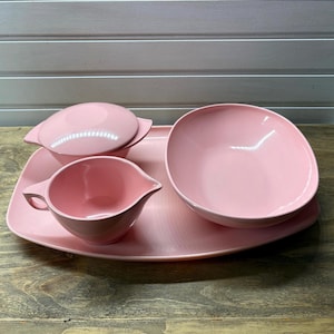 May include: A set of light pink melamine dinnerware consisting of a large rectangular serving tray, a wide shallow bowl, a small lidded container with side handles, and a small creamer pitcher with a handle and a pointed pouring spout.