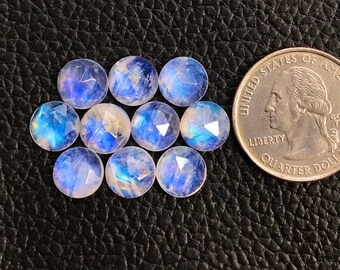 May include: Ten round, faceted moonstone gemstones with blue and white iridescence. A U.S. quarter coin is next to the gemstones for size comparison. The gemstones are arranged on a black textured surface.