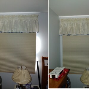 May include: A window featuring a beige pleated cellular shade and a cream-colored ruffled valance with a thin blue trim along the bottom edge. The window is positioned above a wooden nightstand with a lamp and a box of window shade hardware.