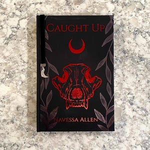 Può includere: Un libro nero con il titolo "CAUGHT UP" in lettere rosse, con una luna crescente rossa e un disegno a teschio. Il nome dell'autore, "NAVESSA ALLEN", è stampato in basso. Un segnalibro in velluto nero con un ciondolo a forma di luna argentata è attaccato.