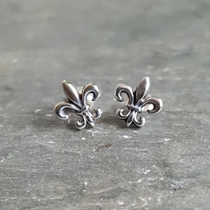 Op de afbeelding: Een paar zilverkleurige fleur-de-lis oorstekers. Elke oorbel heeft een gestileerd ontwerp van de heraldische lelie, een symbool van de Franse monarchie. De oorbellen worden gepresenteerd op een grijs, getextureerd oppervlak.