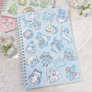 May include: A blue spiral bound notebook with a variety of blue and white cartoon characters on the cover. The characters are all different shapes and sizes and have different expressions on their faces.