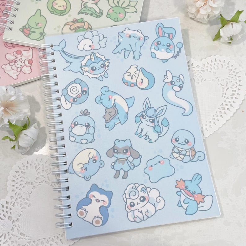 May include: A blue spiral bound notebook with a variety of blue and white cartoon characters on the cover. The characters are all different shapes and sizes and have different expressions on their faces.
