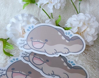 May include: A stack of stickers featuring a cartoon platypus character in shades of brown, gray, and pink. The platypus has a smiling face and the text "TAP TAP" is printed on the sticker. White flowers are in the background.
