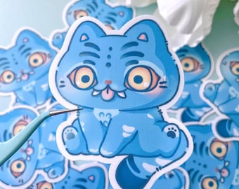 May include: A selection of blue cat stickers with a distinctive design. Each sticker depicts a cartoon cat with large yellow eyes, a pink tongue, and a stylized skeletal pattern. The stickers are on a light blue surface, with tweezers holding one.
