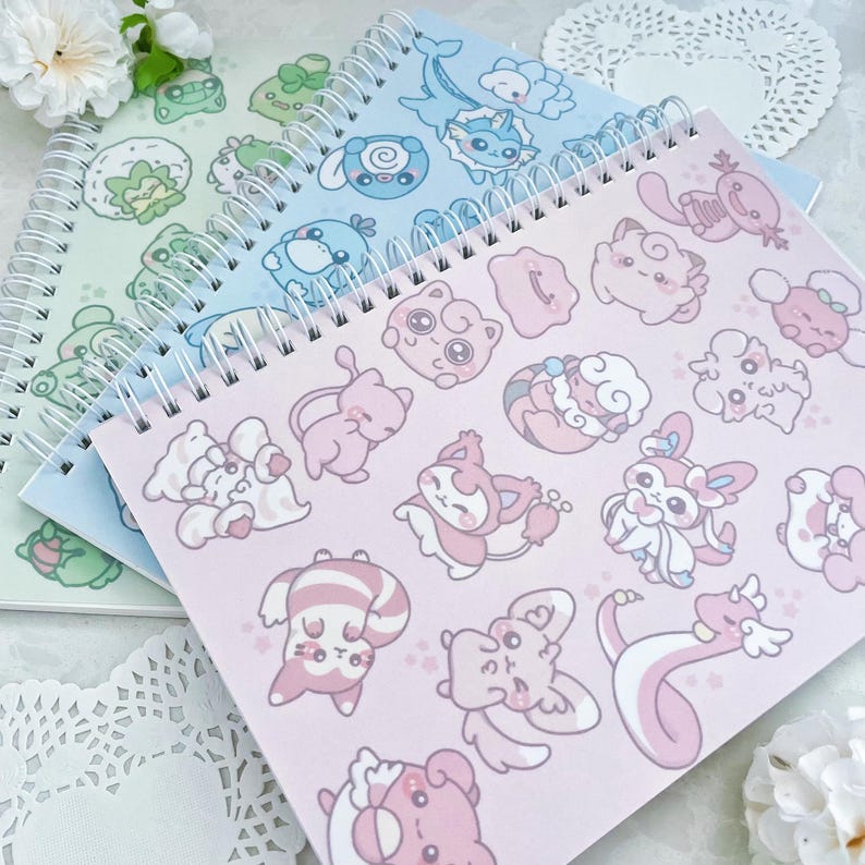May include: Three spiral bound notebooks with different pastel colored covers and cartoon character designs. The covers are light blue, pink, and green. The designs include a variety of cartoon characters, including a blue and white fish, a pink and white cat, and a green and white frog.