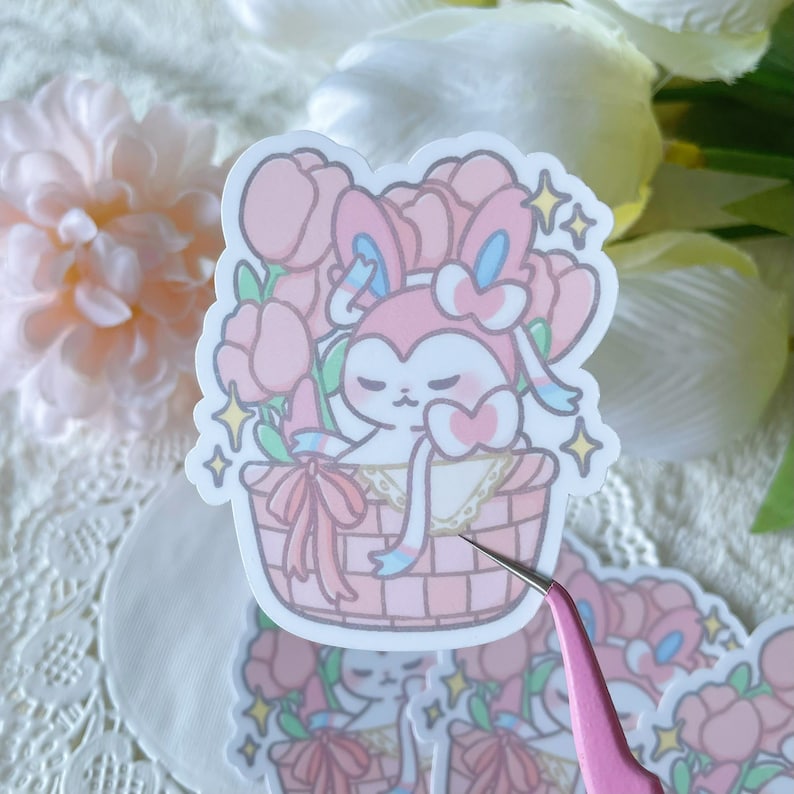 May include: A pink and blue cartoon character with large ears and a bow is sitting in a pink and white basket filled with pink flowers. The character has closed eyes and a pink heart on its chest. The basket is decorated with a pink ribbon and white lace.