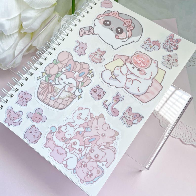May include: A pink and white notebook with a variety of cute cartoon animal stickers. The stickers feature a variety of animals, including cats, dogs, and birds. The stickers are arranged in a random pattern on the notebook.