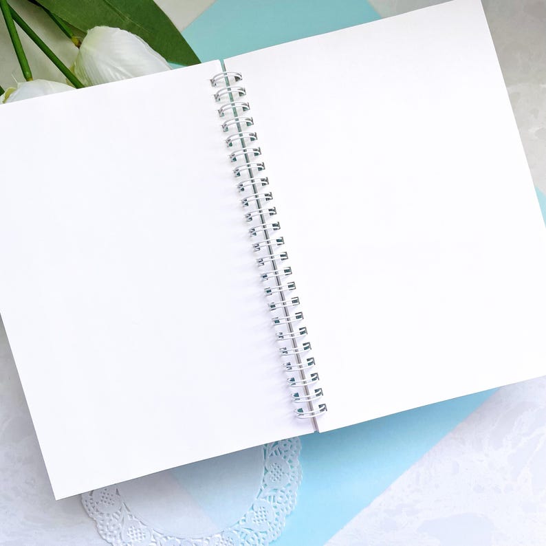 May include: A white spiral bound notebook with blank pages. The notebook is open and lying flat on a light blue surface.