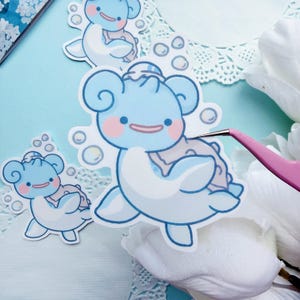 May include: Several stickers featuring a cartoon character in shades of blue and white, with pink cheeks and a smiling expression. The stickers are surrounded by bubbles and appear to be on a light blue surface, with a pair of pink tweezers holding one.