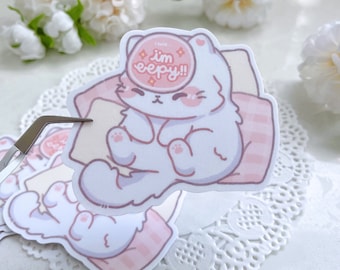 May include: A white cat sticker with pink accents, sleeping on a pink and white striped pillow. The cat has a pink circle over its eyes with the text "I'm sleepy!!" in white.