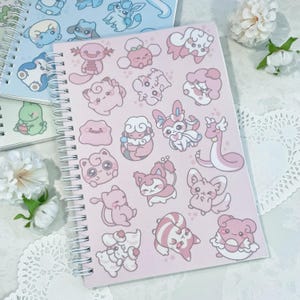 May include: A pink spiral bound notebook with a variety of pastel pink and white cartoon characters on the cover. The characters are all different and include a variety of animals and creatures.