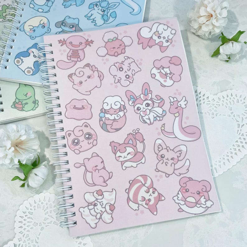 May include: A pink spiral bound notebook with a variety of pastel pink and white cartoon characters on the cover. The characters are all different and include a variety of animals and creatures.