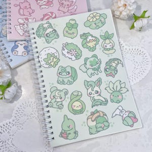 May include: A green spiral bound notebook with a variety of cute cartoon characters on the cover. The characters are green, white, and light pink and include a variety of plants and animals.
