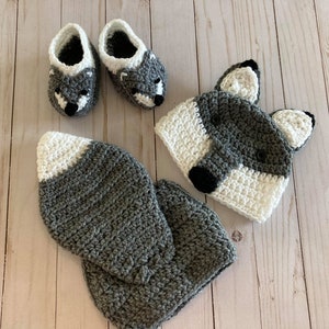 May include: A gray and white crocheted wolf hat and booties set. The hat has two floppy ears and a black nose. The booties have a gray and white pattern and a black nose.