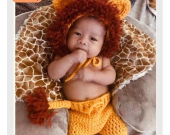 Op de afbeelding: Een baby in een gehaakt leeuwenkostuum, met een bruine en oranje manen, oren en bijpassende broek. De baby ligt op een deken met giraffenprint en een grijs kussen.