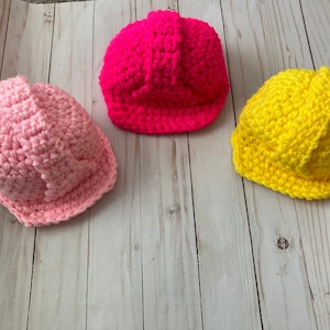 May include: Three crocheted construction worker hats in pink, yellow, and hot pink. The hats are made with a textured stitch and have a brim.