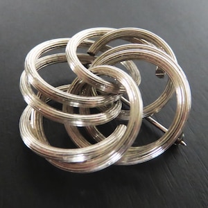 May include: Silver-toned brooch with a complex, interwoven design. The brooch features multiple interlocking circular shapes, creating a three-dimensional, floral-like pattern. The metal has a textured, ridged surface, and a pin closure is visible on the back.