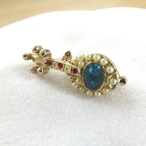 May include: A gold-toned brooch in the shape of a lute. The brooch features a central oval blue gemstone surrounded by small pearls. Red gemstones accent the neck of the lute.