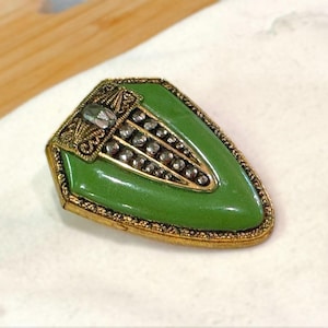 May include: A vintage brooch with a shield shape, featuring a vibrant green enamel center and a gold-toned filigree border. The brooch is adorned with a row of small, sparkling stones and a larger central stone, creating a classic, elegant design.