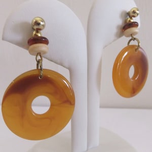 May include: A pair of dangle earrings featuring a gold-tone ball stud, a dark brown disc, a cream-colored disc, and a large, circular, amber-colored resin pendant with a marbled pattern. The components are connected by gold-tone metal rings.
