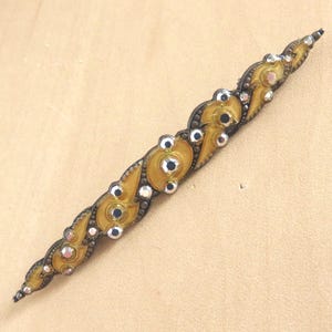 May include: An antique barrette with a decorative design. The barrette has a gold-toned metal base with ornate, swirling patterns. It is embellished with clear and black rhinestones, and has a clasp on the back.