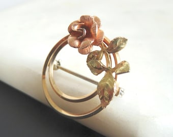Peut inclure: Une broche circulaire ornée d'une rose et de feuilles dans des tons d'or rose et jaune. Le design se compose d'un cadre circulaire fin et lisse en or, avec une fleur de rose texturée et trois feuilles détaillées fixées sur le côté.