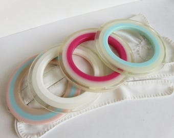 May include: Four colorful, translucent plastic bangles. The bangles feature pink, blue, and white accents. The bangles are stacked on top of a white fabric.