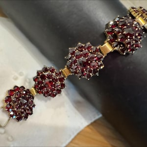 May include: A bracelet featuring four circular clusters of deep red faceted garnet gemstones set in gold-toned metal. Each cluster is connected by small gold-toned metal links, and the bracelet includes a rectangular metal clasp at one end.