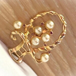 May include: A gold-toned brooch featuring a floral design with faux pearls and small red accents. The brooch has a twisted rope-like border and a decorative openwork element.