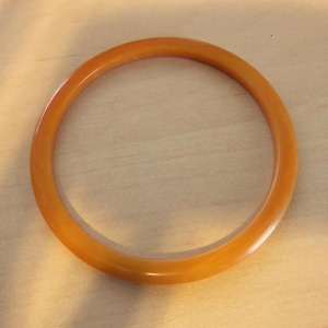 May include: A round, amber-colored bangle bracelet. The bracelet is made of a solid, smooth material and has a glossy finish. The bangle is a classic accessory, suitable for everyday wear or special occasions.