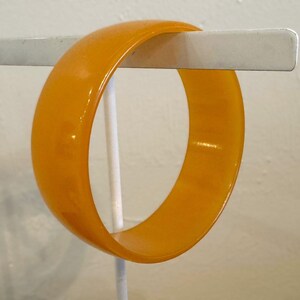 May include: A solid, smooth, mustard-yellow bangle bracelet with a rounded exterior surface and a glossy finish. The circular accessory features a uniform thickness and a consistent, vibrant golden-yellow hue throughout its entire structure.
