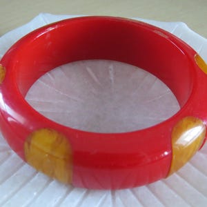 May include: A vibrant red bangle bracelet with three sections of yellow-orange accents. The bracelet is circular and appears to be made of a glossy material. The design is simple and eye-catching, suitable for everyday wear.