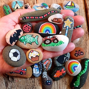 Pu&ograve; includere: Una collezione di sassi dipinti a mano con vari disegni, tra cui un arcobaleno, un serpente, una coccinella e un pallone da calcio. I sassi sono tenuti in una mano, mostrando le colorate opere d'arte su una superficie di pietra naturale.