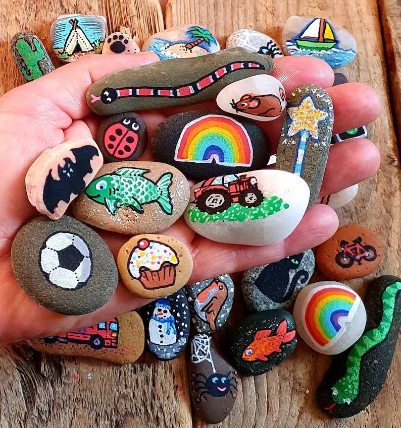 Pu&ograve; includere: Una collezione di sassi dipinti a mano con vari disegni, tra cui un arcobaleno, un serpente, una coccinella e un pallone da calcio. I sassi sono tenuti in una mano, mostrando le colorate opere d'arte su una superficie di pietra naturale.