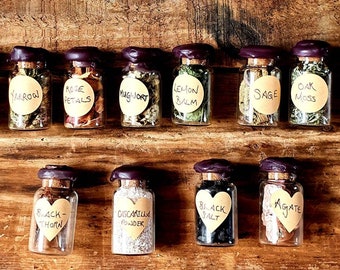 May include: A collection of small glass bottles filled with various herbs and botanicals, each labeled with a handwritten name on a round sticker. The bottles are sealed with corks and dark purple wax. The labels include "Rosemary," "Yarrow," and "Rose Petals."