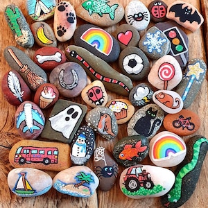 Pu&ograve; includere: Una collezione di sassi dipinti a mano con vari disegni. I sassi presentano immagini di un arcobaleno, un serpente, una coccinella, un gatto, un pupazzo di neve, un autobus, una barca a vela e altre illustrazioni fantasiose.