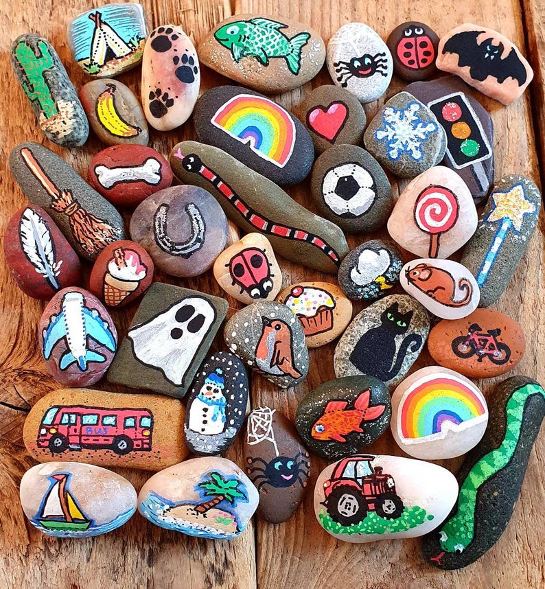 Pu&ograve; includere: Una collezione di sassi dipinti a mano con vari disegni. I sassi presentano immagini di un arcobaleno, un serpente, una coccinella, un gatto, un pupazzo di neve, un autobus, una barca a vela e altre illustrazioni fantasiose.