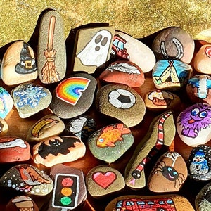 Pu&ograve; includere: Una collezione di sassi dipinti con vari disegni. I sassi presentano immagini di un cappello da strega, un arcobaleno, un fantasma, un pallone da calcio, un ragno, un pupazzo di neve, un cuore e altre illustrazioni colorate. I sassi sono disposti su una superficie di legno.