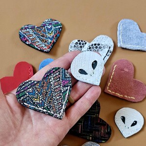 Pode incluir: Uma coleção de patches em forma de coração em várias cores e padrões. Alguns patches apresentam desenhos geométricos coloridos, enquanto outros são vermelhos ou castanhos lisos. Dois patches têm um design de rosto alienígena. Os patches são feitos de tecido e couro.