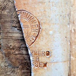 Pode incluir: Close-up de um pedaço de cerâmica quebrado com texto em relevo e um emblema circular. O texto inclui as palavras "BRUNNEN" e "ZBERG". A cerâmica tem uma cor castanha clara com uma superfície áspera e texturizada.
