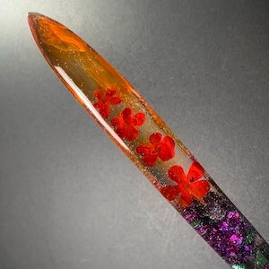 May include: A resin pen with red flowers embedded in a clear, elongated form. The top of the pen has an orange and gold gradient, leading to purple and green glitter at the base. A distinctive writing implement.