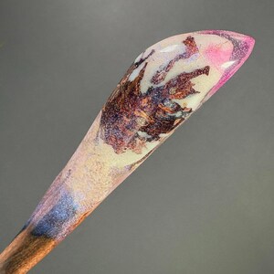 May include: A handcrafted wooden hair stick with a resin top. The resin is a mix of pink, white, and brown, with glitter accents. The wooden base is a dark brown color.